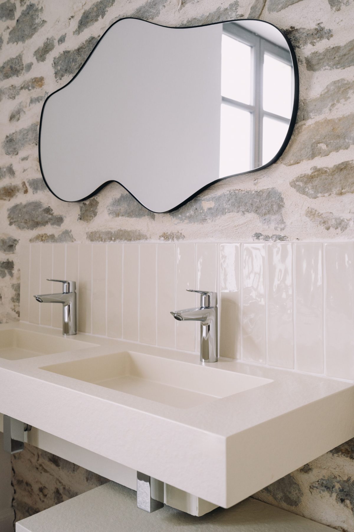 Stylish bathroom interior design with modern fixtures and unique mirror Featuring a contemporary bathroom setup with dual sinks, sleek faucets, and a uniquely shaped mirror against a stone wall, creating a fresh and inviting atmosphere in a residential space.